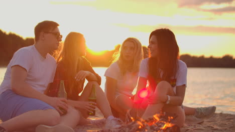 Young-students-are-sitting-around-bonfire-on-the-beach-with-beer.-They-are-talking-to-each-other-at-sunset-and-enjoying-the-summer-evening-on-the-river-coast.